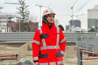 Woman with headscarf under white construction helmet and orange STRABAG clothing stands on the construction site
