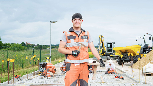 Strabag employee with workwear standing on a street that is being build. In the background are colleagues working and machinery.
