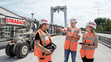 Drei Personen in Schutzausrüstung stehen auf einer Baustelle und unterhalten sich.