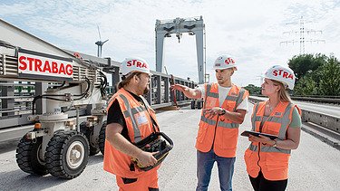 Drei Personen in Schutzausrüstung stehen auf einer Baustelle und unterhalten sich.