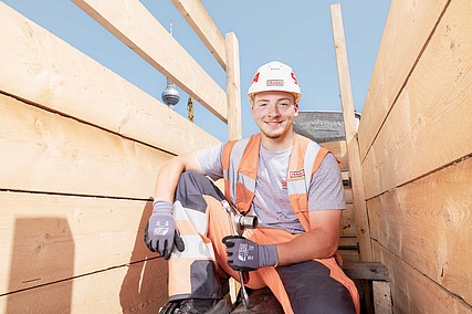 Auszubildender in Arbeitskleidung hockt auf einem Metallrohr