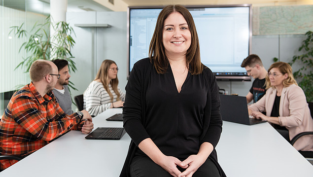 Woman sitting on a desk in a meeting room with people and a presentation screen in the background.          