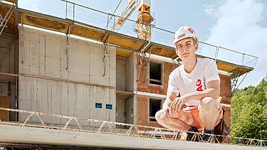 Photo: An apprentice sits in a squatting position on a structural element and looks into the camera