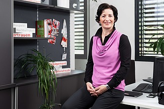 Foto von einer Frau im Büro, die auf der Tischkante sitzt und in die Kamera lächelt