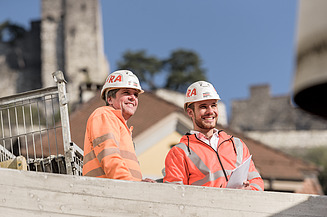 Zwei lächelnde Bauleiter mit Helmen und Warnjacken blicken von einer Baustelle nach oben. Im Hintergrund alte Gebäude.