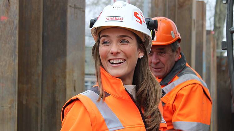 Zwei STRABAG-Mitarbeitende in oranger Sicherheitskleidung auf einer Baustelle; im Vordergrund eine lachende Frau mit weissem Helm mit STRABAG Logo und Gehörschütz, im Hintergrund ein älterer Kollege mit orangen Helm und zwischen Stahlträgern.