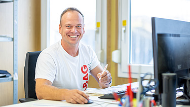 A smiling man sits at a desk and looks into the camera