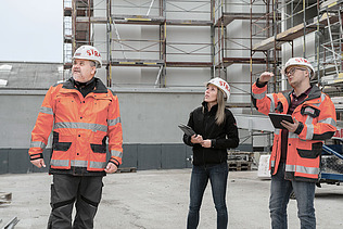 Foto drei Personen auf Baustelle Mann rechts hat Hand angehoben