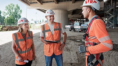 Drei Personen in Arbeitsschutzausrüstung stehen unter einer Brücke und unterhalten sich.