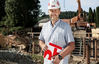 Foto Mann mit Helm und Buch steht vor einer Baugrube