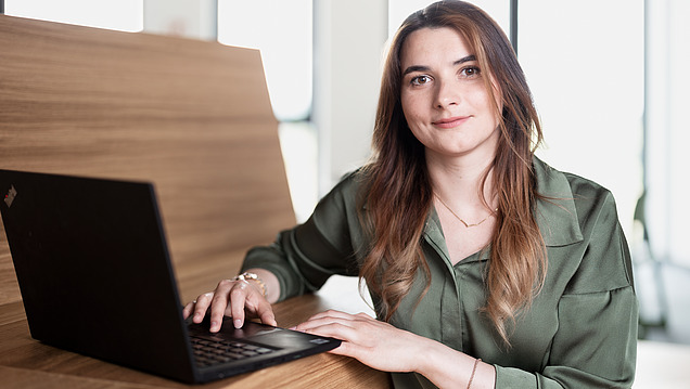 Eine junge Trainee mit braunen Haaren lächelt in die Kamera neben einem Laptop in einem modernen Büro.