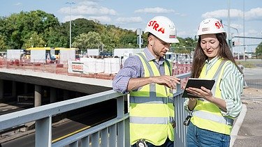 Zdjęcie: Mężczyzna w kasku stoi na placu budowy wraz z Panią Inżynier i monitorują postęp prac za pomocą iPada