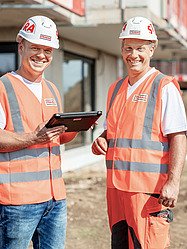 Foto von drei Männern auf der Baustelle, die vor Baugeräten stehen und Arbeitskleidung anhaben