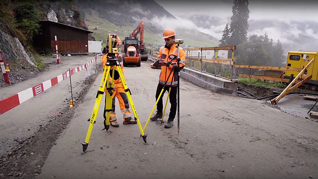 Zwei Bauarbeiter in Warnwesten und Helmen vermessen eine Strasse in den Bergen mit Vermessungsgeräten, auf einer Baustelle. 