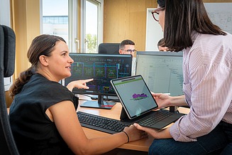 Kolegica sjedi za stolom u uredu, a druga kolegica mu objašnjava i pokazuje nešto na ekranu računala, 