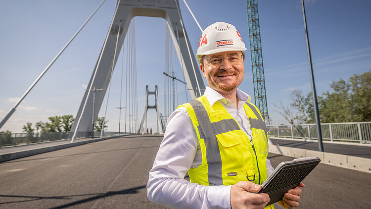 Un bărbat pozează și zâmbește către cameră, cu un pod în fundal.