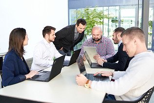 Zdjęcie 6 osób wspólnie siedzi przy stole z otwartymi komputerami i omawiają zadania