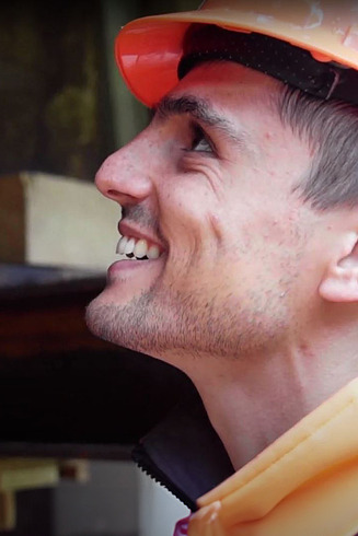 Ein STRABAG-Bauführer lächelt auf seiner Baustelle bei der Arbeit. Er trägt einen Orangenhelm und im Hintergrund kann man Stahlträger verschwommen erkennen. 