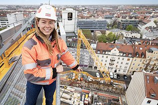 Foto von einer Bauleiterin, die auf einem Bau-Kran steht und auf die Baustelle zeigt