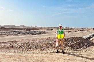 A man in a white construction site helmet and high-visibility vest stands with his hands in his pockets in front of a large construction site in Oman. Sandy earth below, blue sky above.