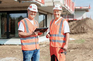 Two construction workers on a construction site. The femal worker is holding a tablet and pointing at something.