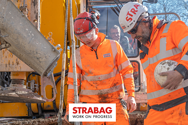 STRABAG Lernender mit weiterem Bauarbeiter in leuchtend oranger Schutzkleidung und Helmen bei der Arbeit an einer grossen Baumaschine – umgeben von Schlamm und Baustellengeräten.