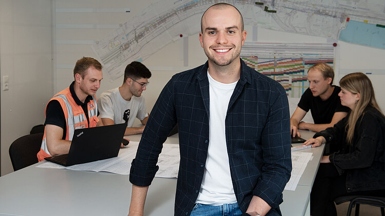 Eine Person sitzt im Vordergrund auf einem Tisch und lächelt in die Kamera, während im Hintergrund vier Personen um den Tisch sitzen.