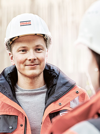 Foto Mitarbeiter mit Bauhelm blickt Mitarbeiterin mit Bauhelm an