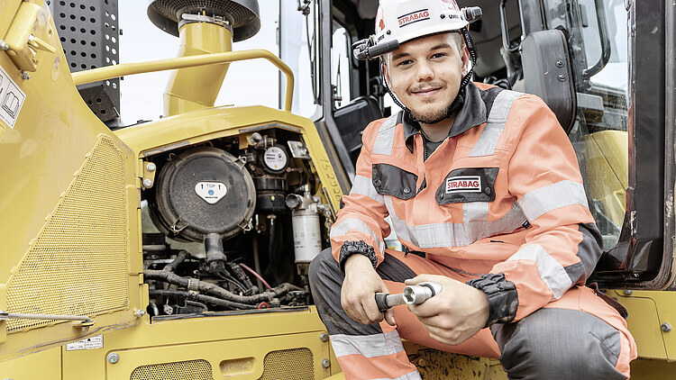 Pripravnik na gradbišču gleda v kamero, za njim je stroj