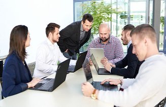 Zdjęcie 6 osób wspólnie siedzi przy stole z otwartymi komputerami i omawiają zadania