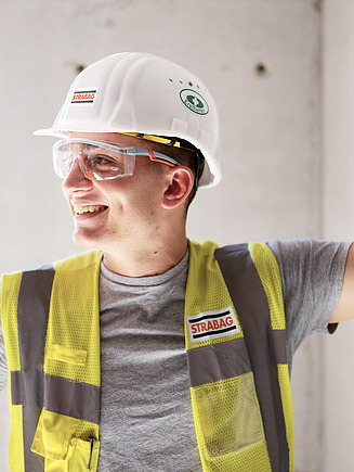 Ein lächelnder Bauarbeiter mit STRABAG-Helm und Schutzbrille trägt eine gelbe Warnweste. Er stützt sich mit einem Arm an der Wand ab auf einer Baustelle.