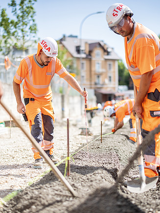 Zwei STRABAG-Bauarbeiter in leuchtender Schutzkleidung befestigen bei Sonnenschein und während dem Betongiessen, Stahlseile an einen Baukran.  Im Hintergrund eine bergige Landschaft mit Bäumen und Häusern.