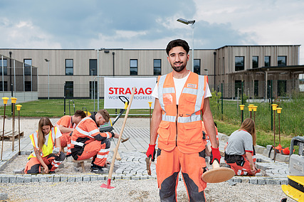 Lehrling in Arbeitskleidung steht im Vordergrund, im Hintergrund pflastern weitere Lehrlinge