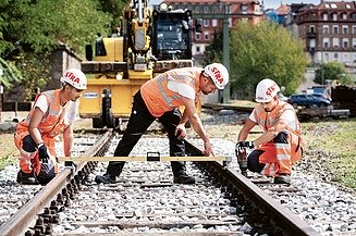 Foto mit 3 Personen die den Abstand von Bahngleisen ausmessen.