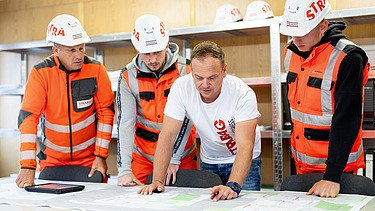 Four men in a portacabin look at a plan on the table