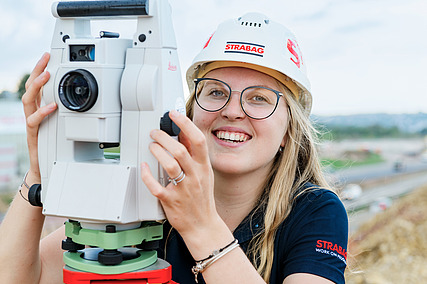 Auszubildende mit STRABAG-Helm steht hinter einem Vermessungsgerät