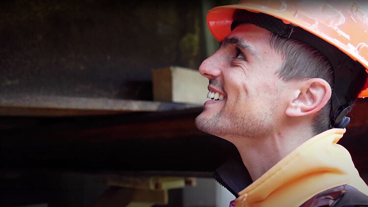 Ein STRABAG-Bauführer lächelt auf seiner Baustelle bei der Arbeit. Er trägt einen Orangenhelm und im Hintergrund kann man Stahlträger verschwommen erkennen. 