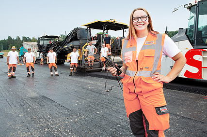 Auszubildende in Arbeitskleidung steht im Vordergrund, während weitere Azubis im Hintergrund arbeiten
