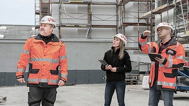 Foto drei Personen auf Baustelle Mann rechts hat Hand angehoben