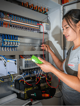 Eine Frau in einem T-Shirt mit STRABAG Logo prüft mit einem Messgerät eine elektrische Schalttafel.