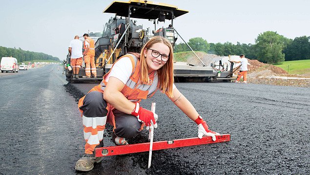 Eine Auszubildende in Arbeitskleidung kniet vor einem frisch asphaltierten Straßenabschnitt.