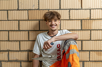 Ein lächelnder Maurer mit Kelle und STRABAG-Arbeitshose sitzt vor einer gemauerten Ziegelwand.