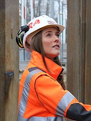 Eine STRABAG Mitarbeiterin lächelt in die Kamera. Im Hintergrund ist ein Lift mit verglasten Türen zu erkennen. 