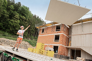 Foto: Ein Bauarbeiter dirigiert eine am Kran hängende Betonplatte.