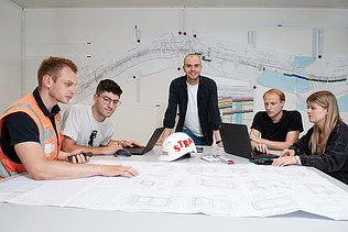 Vier Personen sitzen um einen Tisch herum, während ein Mann an der Mitte des Tisches steht und in die Kamera lächelt.