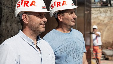 Slika dveh zaposlenih na gradbišču, en zaposleni v ozadju