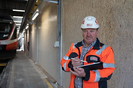 Mann mit Bauhelm auf und Arbeitsjacke an, hält ein Tablet in der Hand und schaut in die Kamera