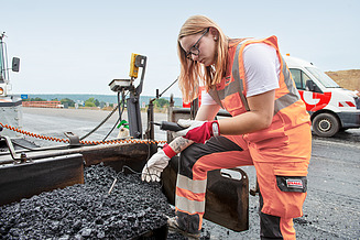 Eine STRABAG Lernende in Schutzkleidung kontrolliert die Temperatur des frischen Asphalts während der Strassenverlegung. Im Hintergrund sind Baustellenfahrzeuge und eine offene Strassenfläche sichtbar.