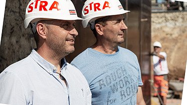 Foto Zwei Mitarbeiter auf Baustelle, ein Mitarbeiter im Hintergrund