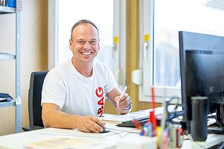 A smiling man sits at a desk and looks into the camera